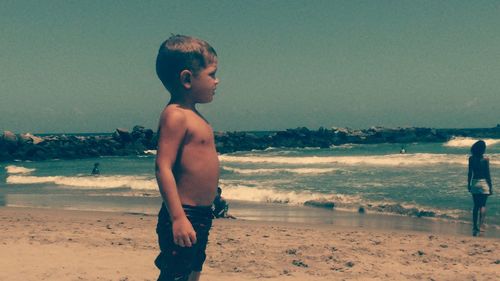 Shirtless boy standing at beach against sky