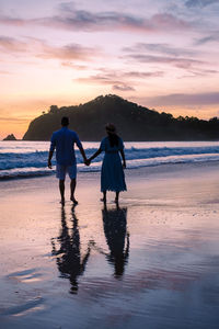 Rear view of man standing at beach against sky during sunset