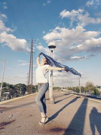 Full length of woman standing on bridge while holding waving scarf against cloudy sky