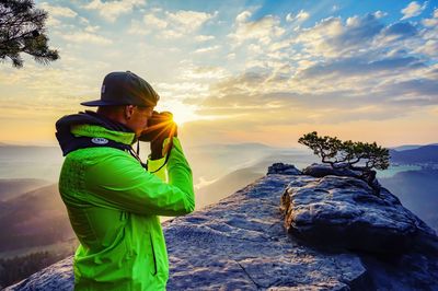Man photographing with camera while standing on mountain against sky during sunset