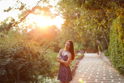 Portrait of mid adult woman with arms crossed standing on footpath amidst plants during sunset