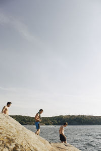 Shirtless male friends walking towards lake against sky at sunny day