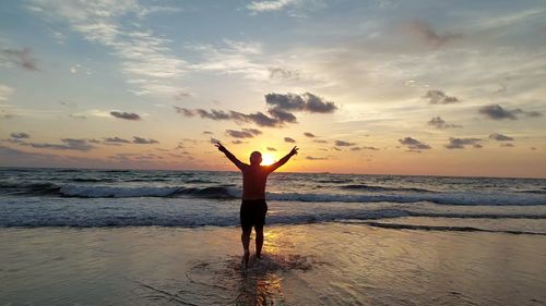 Rear view of man with arms outstretched standing at beach against sky during sunset