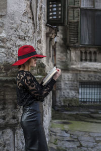 Side view of woman reading book while standing by wall