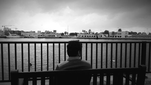 Rear view of man sitting by railing in city against sky