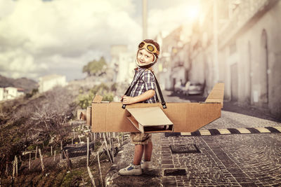Portrait of boy in cardboard airplane on road against sky