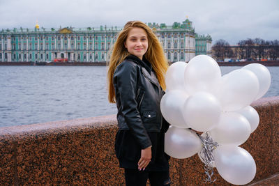 Caucasian girl holding white balloons standing by river on embankment of city