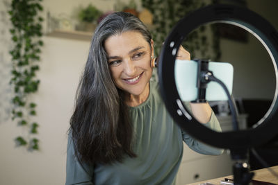 Smiling mature woman applying foundation while filming through smart phone at home