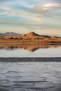 A hill is reflected in a partially frozen lake with ice in the foreground