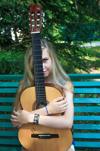 Portrait of young woman holding guitar while sitting in bench at park