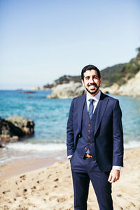 Portrait of smiling bridegroom standing at beach against clear sky during sunny day