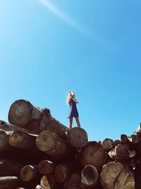 Low angle view of woman standing on logs against clear blue sky