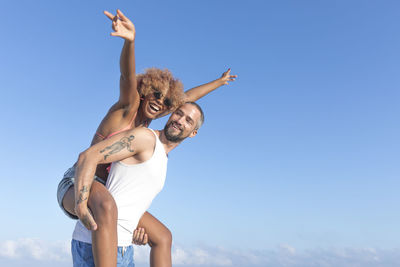 Low angle view of man piggybacking cheerful girlfriend against blue sky