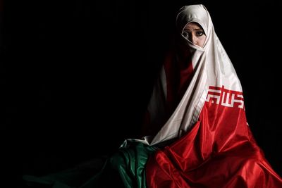 Portrait of woman wrapped in iranian flag against black background