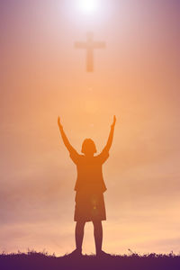Digital composite image of boy with arms raised standing below cross against cloudy sky during sunset