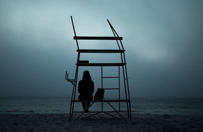 Rear view of silhouette woman sitting on built structure at beach