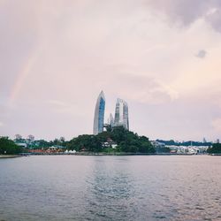 View of city buildings by river against cloudy sky