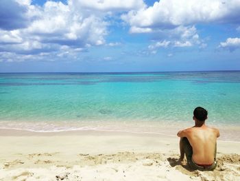 Rear view of shirtless man sitting on sand against sea at beach