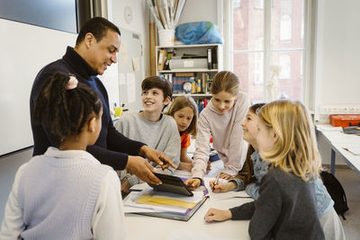 Male teacher teaching students through tablet pc at desk in classroom