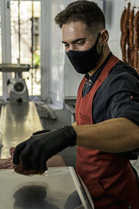 Butcher with mask making raw beef burgers in butchers shop.