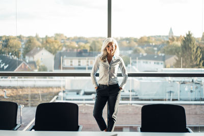 Smiling businessman with hands in pockets standing in front of glass window at work place