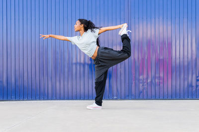 Full body side view of active african american female dancer standing on one leg and holding foot behind back bear blue wall