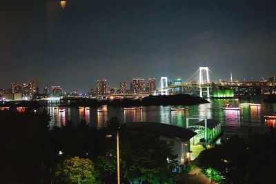 Illuminated modern buildings by river against sky at night