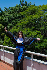 Portrait of smiling young woman standing by railing against trees