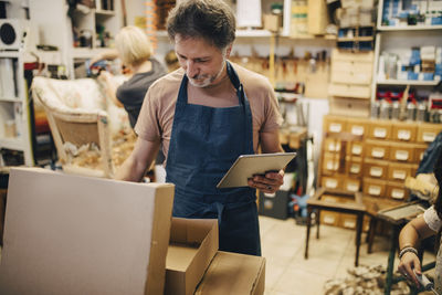 Male craftsperson holding digital tablet while opening cardboard box in workshop