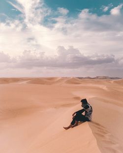 Man sitting on desert against cloudy sky
