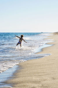 Rear view of shirtless boy enjoying on shore at beach against sky