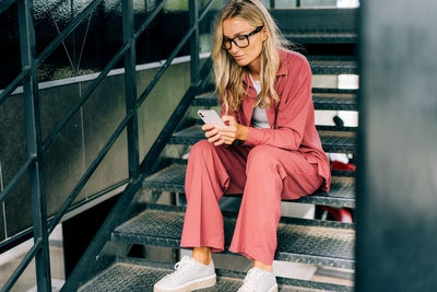 Young casual woman using smartphone while sitting on the iron steps of a modern office building.