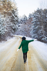 A young girl in a green sweater and hat walks in the middle of a snowy road in a thick pine forest.