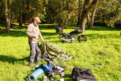 Senior man removing tent from grassy field in forest during sunny day