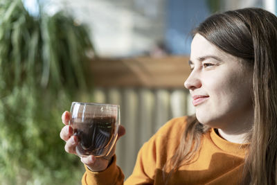 Smiling young pretty caucasian woman holding glass cup of coffee on background of green house plants