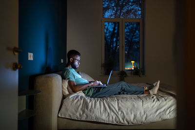 Black student guy lying in bed with laptop in evening, studying until late hours