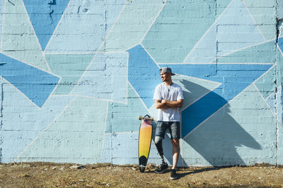 Young man with leg prosthesis standing against concrete wall with a skateboard