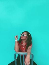 Young woman wearing sunglasses sitting against blue wall