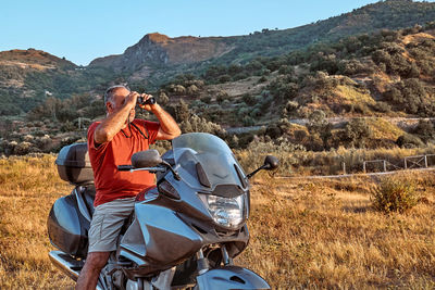On the road.tourist man traveling on motorcycle, looking through binoculars at mountains in sunset