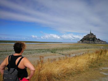 Rear view of mid adult man with backpack looking at castle while standing on field against cloudy sky