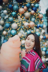 Portrait of smiling young woman standing by christmas tree holding cotton candy outdoors