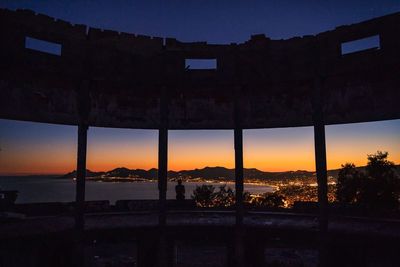 Silhouette buildings by sea against sky during sunset