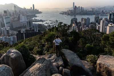 Rear view of man looking at city while standing by rocks against sky during sunset