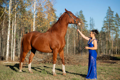 Side view full length of woman standing by horse at forest