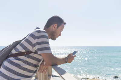 Side view of man using phone while standing at beach against clear sky