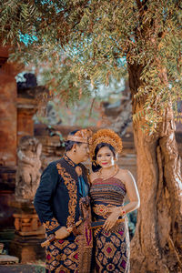 Balinese couple standing against tree wearing balinese traditional clothes
