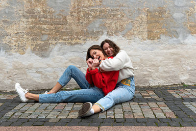 Two best friends hugged sitting on a paved sidewalk by an old wall