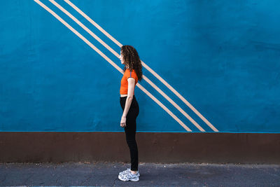 Young woman standing on footpath in front of blue wall