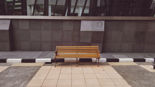 Empty bench by sidewalk against building in city