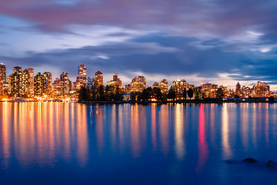 Illuminated buildings by lake against sky in city